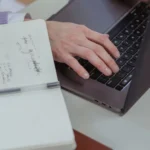 Dłoń pisze na klawiaturze laptopa obok notatnika z odręcznymi notatkami